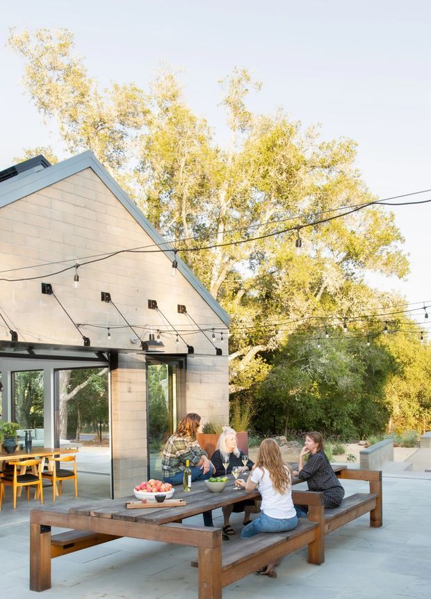A bright outdoor dining and entertaining area beside a contemporary gabled pavilion. The scene features a large timber picnic-style dining table with built-in benches, pendant string lighting suspended across the patio, exposed dark structural beams, pale masonry walls, extensive glazing, and a landscaped garden setting. A dining room is visible through the open glazed doors.
