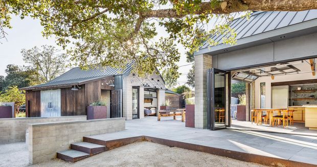 A contemporary single-storey residential courtyard and outdoor entertaining area. The composition shows a main pavilion with a broad glazed opening, pale masonry walls, dark metal standing-seam roof and an open-plan kitchen/dining space inside. A smaller dark timber-clad pavilion sits beyond the concrete courtyard, with low concrete retaining walls, rust-toned planter boxes and steps. Mature trees frame the scene.