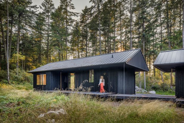A compact dark-clad cabin set within a dense forested landscape. The single-storey building has a simple pitched metal roof, vertical dark timber-like cladding, a covered entry or deck, and warm-lit windows. A second matching structure is partially visible on the right, connected by a dark walkway or deck.