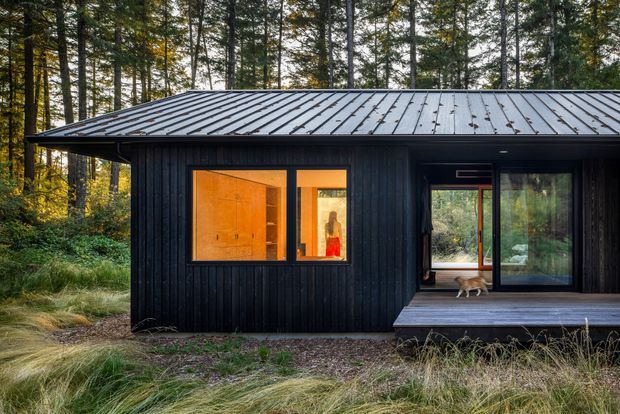 A compact single-storey black-clad cabin set within a wooded landscape. The building has a low-pitched standing-seam metal roof, vertical dark timber-like cladding, warm illuminated windows, and a glazed sliding-door opening onto a raised timber deck. Forest vegetation surrounds the structure.
