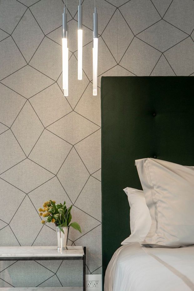 A close-up view of a refined bedroom corner featuring a tall dark green upholstered, button-tufted headboard, crisp white bedding, and a slim console or bedside table with a glass vase of flowers. The wall is covered in a grey textured geometric patterned finish, while three slender pendant lights hang overhead. The composition has a sophisticated, modern feel with strong geometric detailing and a restrained palette of grey, white, black, and deep green.