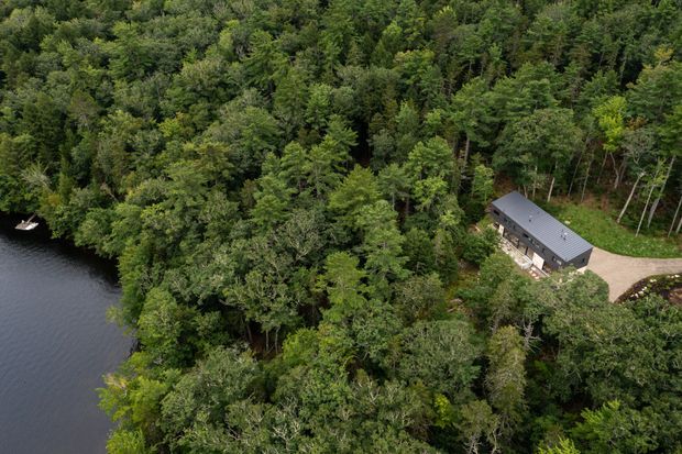 Aerial view of a secluded modern, elongated dark-roofed building nestled among dense forest beside a calm lake. The property includes a pale gravel or paved approach and a small dock or boat visible along the shoreline.