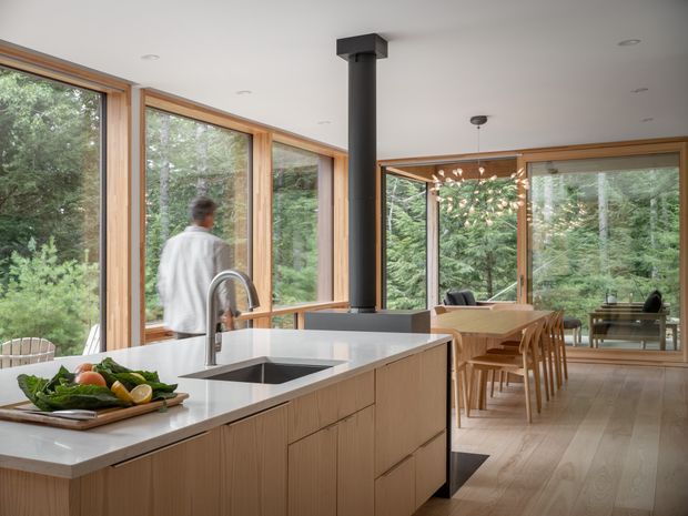 Open-plan kitchen and dining area surrounded by floor-to-ceiling timber-framed glazing overlooking a wooded landscape. The kitchen features a large light-wood island with a pale stone benchtop, undermount sink and brushed metal tap, while a central dark cylindrical fireplace or flue creates a strong focal point. A timber dining table and chairs sit beyond the island, with warm timber flooring, recessed ceiling lights and a sculptural pendant. The space is bright, calm and strongly connected to nature.