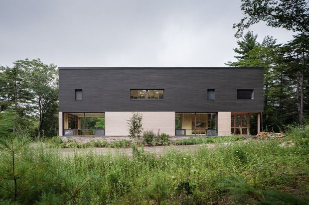 Front exterior view of a long, rectilinear two-storey house set within dense natural woodland and meadow planting. The upper level is clad in dark horizontal timber or composite boards with small recessed windows, while the lower level combines pale masonry or rendered panels with extensive timber-framed glazing. The design has a minimalist contemporary form and a flat roofline.