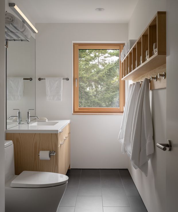 A compact, bright bathroom with a wall-mounted timber vanity, white basin, toilet, large mirror, dark grey tiled floor, and extensive open timber shelving with towel hooks. A timber-framed window provides natural light and views of trees. The design is clean and contemporary with warm wood accents and practical built-in storage.