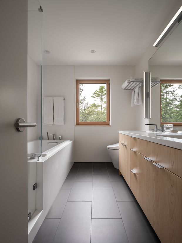 A bright, contemporary bathroom viewed from the doorway, with a freestanding-style white bathtub enclosed by a clear glass shower screen on the left, a toilet at the rear, and a long timber-veneer vanity with a white countertop and integrated sink on the right. Large mirrors, a wall-mounted towel rack, recessed ceiling lights, and a linear light add functionality. The room has dark grey tiled flooring, pale painted walls, warm timber window frames, and views of trees through the windows.