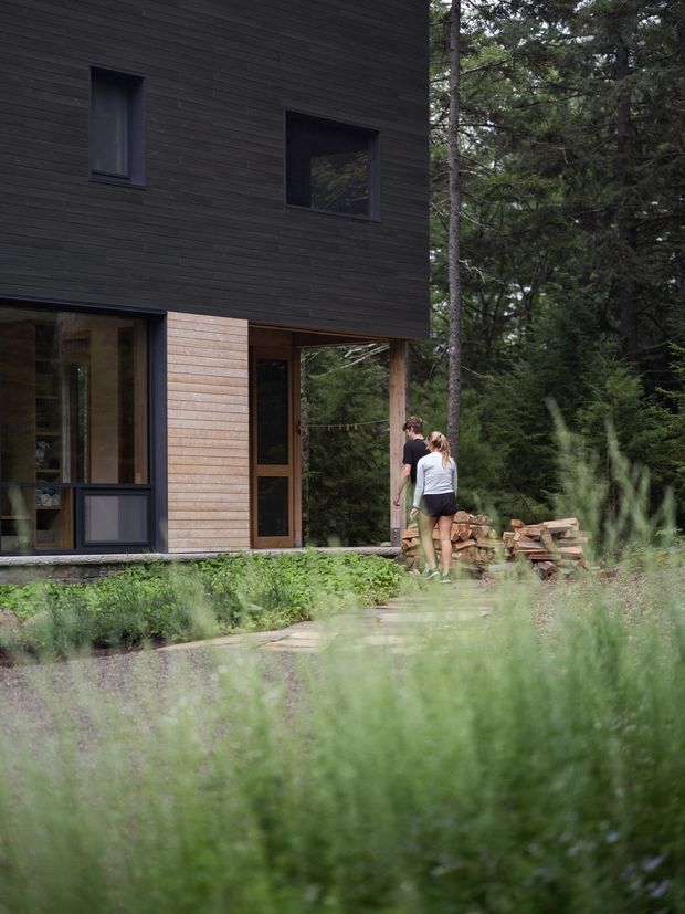 Exterior view of a compact two-storey house set within a dense forest. The building has dark horizontal timber cladding, large dark-framed windows, a lighter timber-clad entry volume, and a partially recessed ground floor supported by timber posts. A path and rough stacked timber sit in the landscaped foreground, with two people near the entrance.