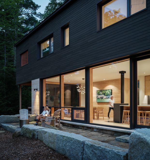 Exterior dusk view of a contemporary two-storey house set among trees, with a dark horizontal-clad upper facade and extensive warm timber-framed ground-floor glazing. The open-plan interior is visible through the windows, including a kitchen or dining area with timber walls and stools. Large rough stone blocks form a low landscape edge in the foreground, with outdoor seating beside the house.