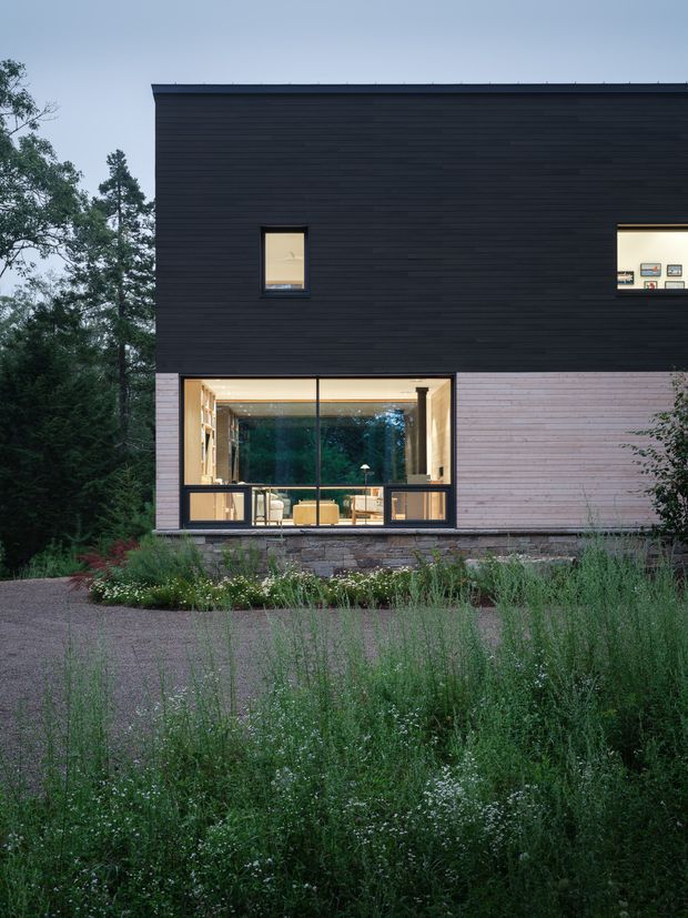 A contemporary two-storey house viewed from the landscaped front garden. The facade combines dark horizontal cladding on the upper level with pale horizontal cladding and a low stone base below. Large black-framed glazed openings reveal a warmly lit interior, while smaller upper windows punctuate the simple rectangular form. Dense planting, gravel paving, and mature trees surround the house.