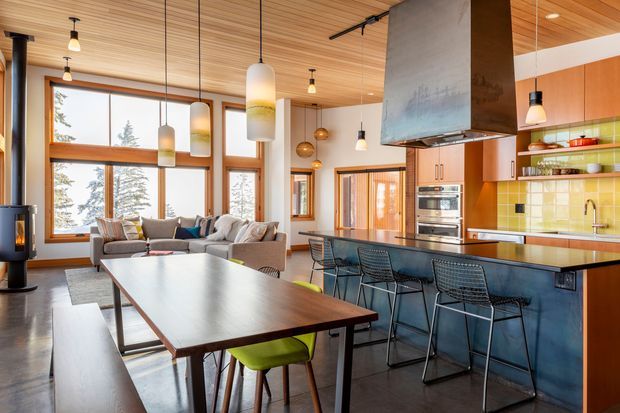 Open-plan contemporary kitchen, dining and living area with a warm timber slatted ceiling, expansive timber-framed windows, dark flooring, and a large island with blue cabinetry and a bright yellow tiled splashback. A dining table occupies the foreground, while a sectional sofa and freestanding wood-burning stove sit near the windows. Pendant lights, exposed kitchen shelving, and a large sculptural rangehood add visual interest.