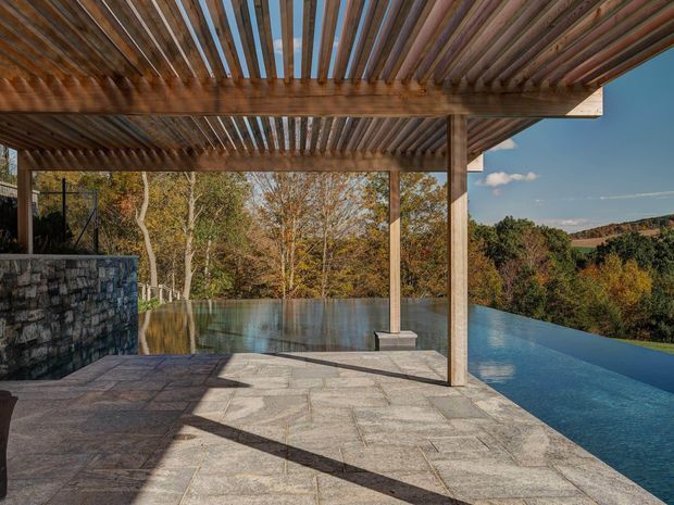 An outdoor pool terrace beneath a substantial timber pergola. The terrace is paved with large irregular natural stone slabs, with a dark stacked-stone wall at left and slender timber posts supporting the overhead slatted roof. An infinity-edge pool extends toward a wooded, hilly landscape, creating a strong connection between the built outdoor space and its natural setting.