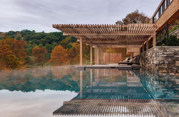 Exterior architectural view of a timber-clad residence beside a reflective swimming pool, featuring a large timber pergola extending over the pool terrace, stone retaining walls, dark-framed glazing, and a wooded landscape beyond. The image has a calm, refined, nature-integrated character with strong reflections in the still water.
