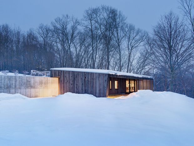 A low, minimalist single-storey building set in a snowy wooded landscape. The long rectangular form has vertical weathered timber cladding, a shallow flat or very low-pitched roof, and warm recessed glazing and doorway lighting. A smooth concrete or rendered wall projects at the left, while deep snow covers the foreground and surrounding site.
