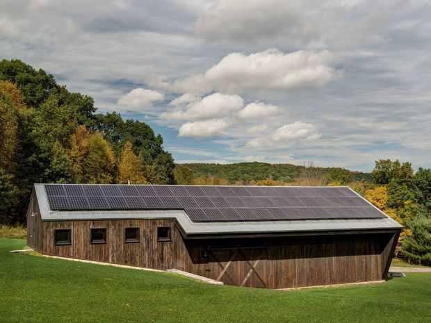 A low, elongated timber-clad barn-like building set in a grassy rural landscape, with a broad pitched roof covered in solar panels, simple rectangular windows, and large dark timber doors. Dense tree-covered hills and a cloudy sky form the background.