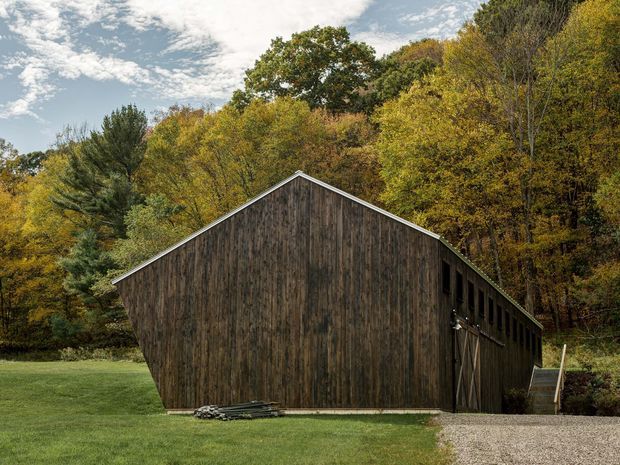 Exterior view of a simple barn-like timber building set in a wooded, rural landscape. The structure has a steep central gable roof, vertical dark weathered timber cladding, a large barn door, and a gravel approach, with autumn trees surrounding the grassy site.