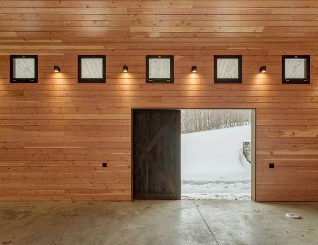 Interior view of a timber-clad garage or outbuilding with horizontal wood wall and ceiling lining, a polished concrete floor, small square windows, warm wall lights, and a large dark timber barn-style door opening onto a snowy exterior.