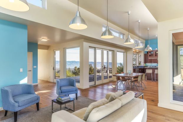 Bright open-plan living, dining and kitchen area with a warm timber floor, white walls and ceiling, a vivid blue accent wall, multiple pendant lights, and extensive glazed doors and clerestory windows overlooking a scenic landscape. The room contains a large light sofa, blue lounge chairs, a coffee table, dining table, and a compact kitchen with dark red-brown cabinetry.