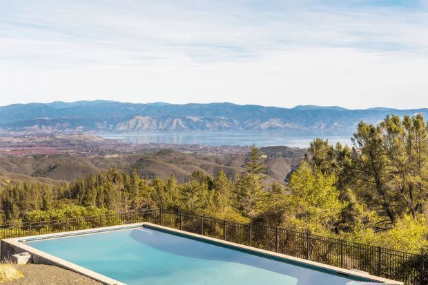 An elevated outdoor swimming pool overlooks a broad mountainous landscape and a large body of water, with wooded hills and a distant shoreline visible beyond the pool terrace. The scene is bright and expansive, with black metal fencing and pale pool coping framing the foreground.