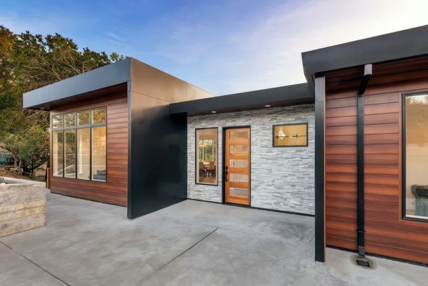 Exterior view of a contemporary single-storey residence with a low-slung, asymmetrical flat-roof form. The facade combines horizontal warm timber cladding, dark metal fascia and structural elements, and a central grey stacked-stone wall around the timber entry door. Large glazed openings and a broad concrete forecourt reinforce the modern indoor-outdoor character, with mature trees visible beyond.