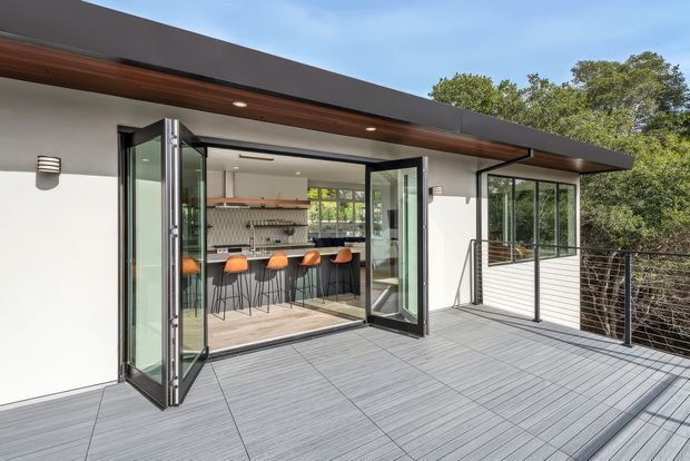 Exterior view of a contemporary flat-roof home opening onto a spacious grey deck terrace. Large black-framed folding glass doors connect the terrace to a bright kitchen with a long island, bar seating, timber cabinetry, and a patterned backsplash. The facade combines white rendered walls, dark roof trim, timber soffit detailing, and slim metal cable balustrades.