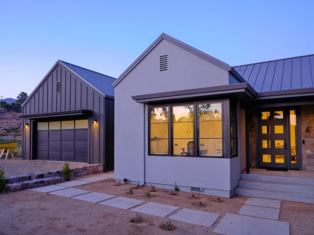 Exterior view of a contemporary single-storey home at dusk, featuring a gabled central entrance volume, dark standing-seam metal roofs, pale rendered walls, a large glazed bay window, and a dark garage wing with a sectional garage door. The entry includes a glazed dark-framed door beside a stone-clad wall, with concrete stepping paths and sparse drought-tolerant landscaping.