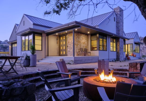 Twilight view of a low-rise contemporary home with multiple pitched metal roof sections, pale rendered walls, large dark-framed windows, and a prominent natural-stone chimney. A covered entry porch opens toward a landscaped outdoor seating area with Adirondack-style chairs and a round fire pit.