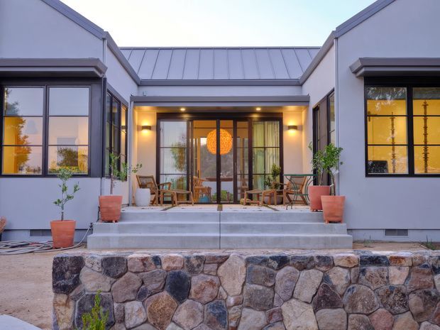 Front-facing exterior view of a contemporary single-storey home with a symmetrical courtyard-like entry, broad concrete steps, dark-framed glazing, a standing-seam metal roof, and a low natural stone retaining wall. Warm interior lighting and an orange pendant are visible through the central glazed entrance, with potted plants and outdoor seating on the entry terrace.