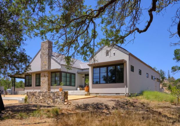 Exterior view of a contemporary single-storey home set in a dry, lightly wooded landscape. The house has pale rendered walls, a prominent natural stone chimney and low retaining wall, dark-framed glazing, a light metal gabled roof, and covered outdoor areas. Mature tree branches frame the view, with dry grasses and rocky ground surrounding the building.
