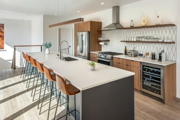 Bright, spacious contemporary kitchen with a large waterfall-edge island and multiple leather bar stools. The room features warm timber cabinetry, stainless steel appliances, a geometric patterned tile splashback with open timber shelves, and light wood flooring. A stair landing is visible beyond the kitchen, with abundant white surfaces and recessed lighting creating an airy, refined atmosphere.