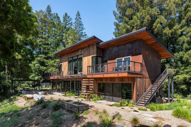 Exterior view of a contemporary two-storey timber-clad house set among mature trees. The home has a strong horizontal composition with offset low-pitched roof planes, extensive glazing, a projecting upper-level balcony, and an external metal stair leading to the deck. A landscaped gravel and planting area surrounds the building.