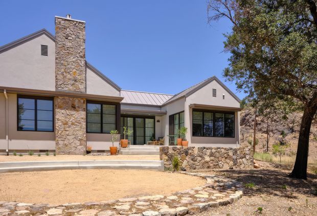 Single-storey residential house viewed from the front, with pale rendered gabled wings, a standing-seam metal roof, large dark-framed windows and doors, and prominent irregular natural-stone chimney and retaining wall elements. The house sits in a dry, lightly landscaped setting with mature trees and a broad sandy forecourt.
