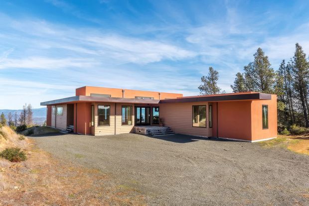 Single-storey modern residence viewed from the front across a broad gravel forecourt. The house has a low, horizontally composed form with flat roofs, projecting roof planes, warm terracotta-orange rendered wall sections, pale horizontal timber-like cladding, dark window and door frames, and a recessed central entry with steps. Tall trees and distant terrain are visible around the property.