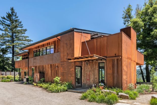 Exterior view of a contemporary two-storey residence with a strongly rectilinear form, extensive warm weathered timber or rust-toned cladding, deep projecting eaves, exposed structural elements, and a partially screened upper-level window band. Landscaping and mature trees surround the entrance, while a gravel driveway or forecourt occupies the foreground.