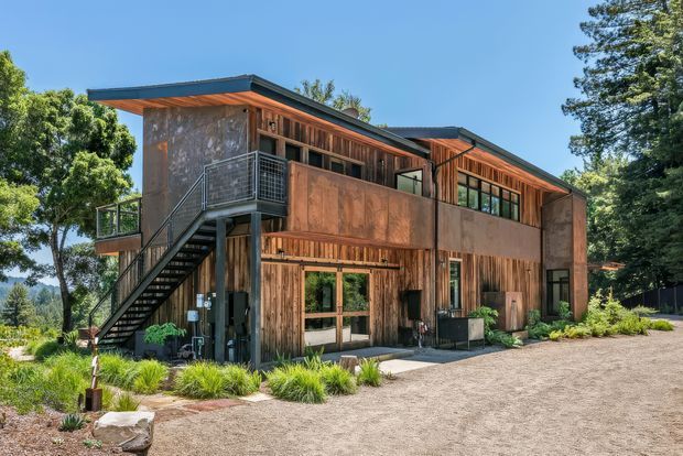 Exterior view of a contemporary two-storey residence set among mature trees. The building features extensive weathered timber cladding, large glazed openings, a low-pitched projecting roof, an exposed external metal stair and upper-level balcony, with landscaped planting and a gravel forecourt.