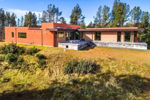 Single-storey modern house set in a wooded, semi-rural landscape. The long, low-slung facade combines terracotta-painted masonry at the left with horizontal timber-toned cladding at the right, dark-framed windows, a shallow roofline, and a raised deck or patio with steps at the entrance.