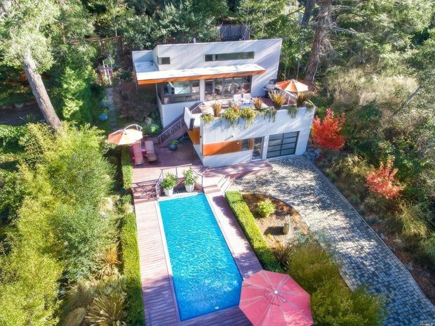 Aerial view of a modern multi-level home integrated into a densely wooded landscape. The house has flat roof forms, extensive glazing, warm timber-toned detailing, planted roof terraces, a long rectangular swimming pool with timber decking, and a paved driveway or courtyard.