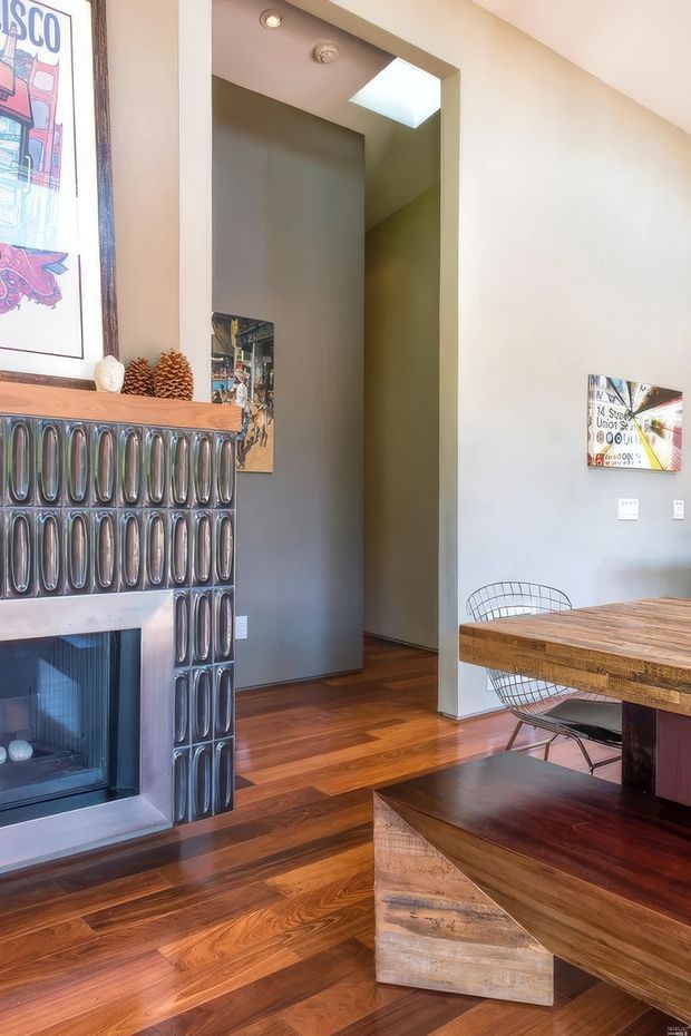 Warm, eclectic open-plan dining and living area with polished timber flooring, a rustic timber dining table and bench, and a distinctive tiled fireplace with a timber mantel. A tall opening leads to a hallway or adjacent space, with a skylight above. Framed artwork and decorative objects add character.