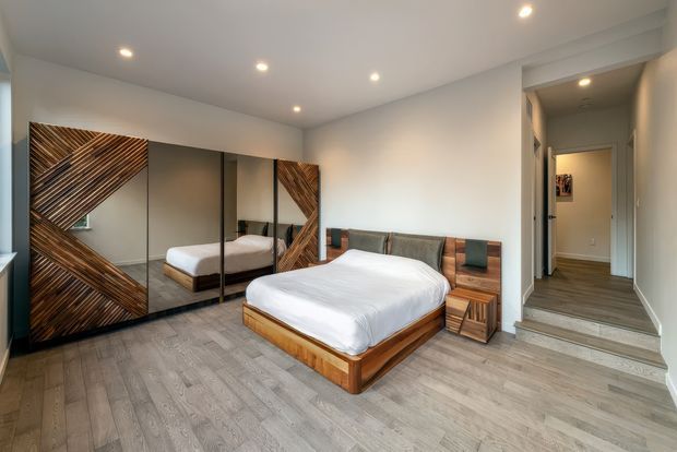 A contemporary bedroom with a low timber platform bed, white bedding, integrated timber bedside tables, and a wood-panelled headboard wall. Large mirrored wardrobe doors reflect the bed and room, while diagonally arranged dark timber wall panels create a geometric feature. The room has pale wood-look flooring, white walls and ceiling, recessed downlights, and a short set of steps leading to a hallway.