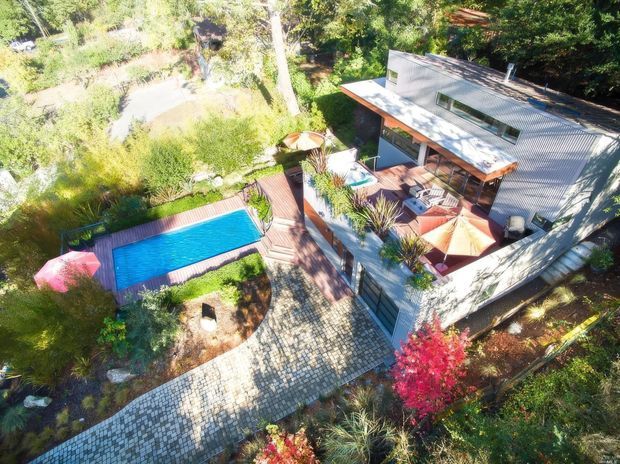 Aerial view of a contemporary multi-level home set within a lush landscaped garden. The property features a corrugated metal roof, extensive timber decking and balconies, a bright rectangular swimming pool, outdoor seating and umbrellas, and a broad paved walkway connecting the house and pool area.