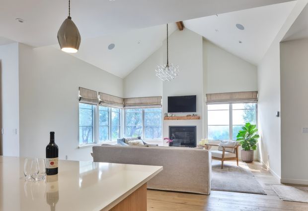 Bright open-plan living area viewed from the kitchen, featuring a vaulted ceiling with exposed timber beam, large windows with woven Roman blinds, a central fireplace with timber mantel and wall-mounted television, pale sofa seating, timber flooring, and a contemporary kitchen island with a light benchtop in the foreground. A sculptural pendant and chandelier provide decorative lighting.