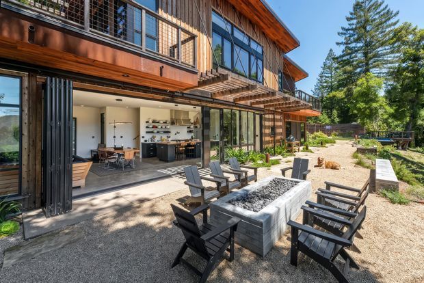 A contemporary multi-level timber-clad residence viewed from a landscaped outdoor entertaining area. Large glazed openings connect the gravel courtyard and seating area to an open-plan kitchen and dining space, while an outdoor rectangular fire feature is surrounded by dark timber lounge chairs. The building has extensive black-framed glazing, projecting timber eaves, upper-level balconies, and a warm natural-material palette.