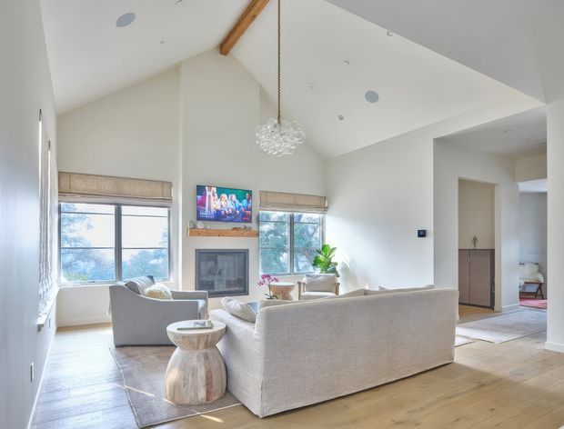 Bright, airy living room with a high sloped ceiling, exposed timber beam, pale walls, light timber flooring, and large windows fitted with woven Roman blinds. A fireplace with timber mantel and wall-mounted television forms the focal point, while a large pale sofa, armchairs, coffee table, rug, and glass pendant create a comfortable contemporary interior. An adjacent hallway and cabinetry are visible beyond the room.
