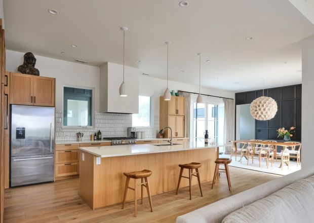 Bright, open-plan contemporary kitchen and dining area viewed from an adjacent living space. The kitchen features a large timber island with a light stone benchtop, timber cabinetry, stainless-steel appliances, a white small-format tiled splashback, pendant lights, and timber flooring. A dining table and chairs sit beyond the island, with dark built-in cabinetry and large glazed doors in the background.