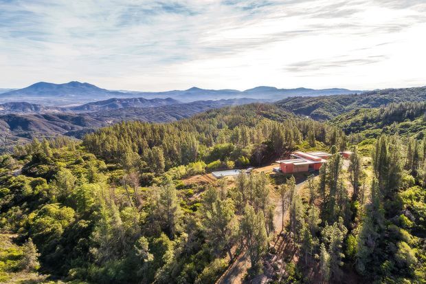Aerial view of a low-profile building with light walls and a red roof nestled among dense green trees on a heavily wooded, sloping landscape. Layered hills and rugged mountain ranges extend across the distant background under a bright, partly cloudy sky.