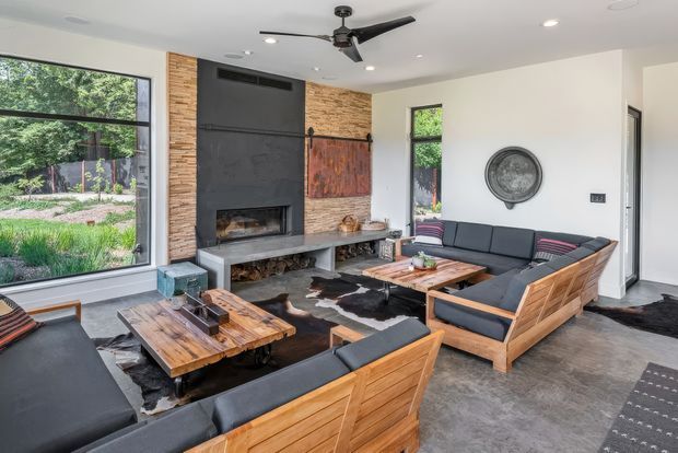 Contemporary living room with expansive glazing overlooking a landscaped garden, a dramatic dark fireplace wall with stone and timber accents, exposed log storage, concrete-look flooring, and low-profile timber-framed lounge seating arranged around rustic wood coffee tables. A ceiling fan and recessed lighting complete the relaxed, modern interior.