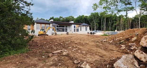 A low, modern single-storey house shown from the front during construction, with broad glazed openings, pale rendered walls, dark grey hipped roof sections, and a covered entry or outdoor living area. The surrounding site is cleared and uneven, with exposed soil, rocks, construction equipment, and forested vegetation beyond.