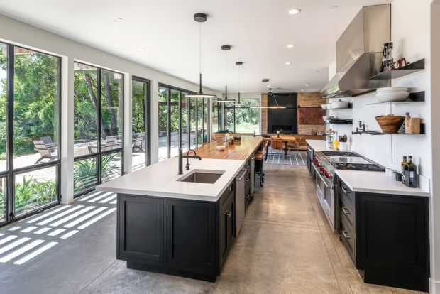A long, contemporary galley-style kitchen with a large central island, dark panelled cabinetry, pale stone benchtops, stainless-steel appliances, open shelving, and extensive floor-to-ceiling glazing along one side. The kitchen opens toward a leafy outdoor setting and includes an integrated dining/work area at the far end. Polished concrete flooring and a restrained black, white, and timber palette create a refined industrial-contemporary feel.