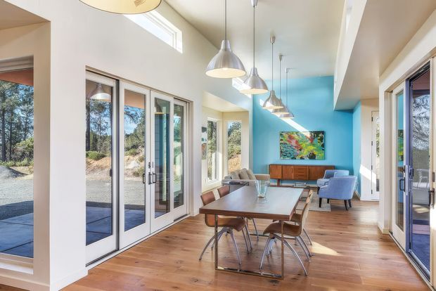 Bright, spacious open-plan dining and living area with warm timber flooring, extensive glazing and multiple glazed doors, a vivid turquoise feature wall, and a row of pendant lights. The dining table and chairs occupy the foreground, with a compact lounge and timber sideboard at the far end. The room has a high, partially vaulted ceiling and strong indoor-outdoor visual connection.