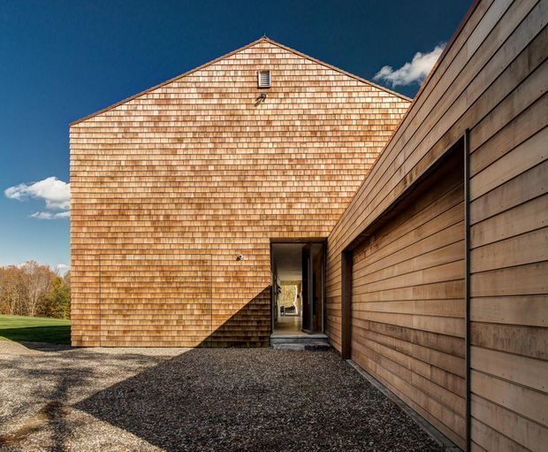 Exterior view of a contemporary, barn-like building clad in warm cedar shingles and horizontal timber boards. A steep gabled facade with a small upper vent faces a gravel approach, while a lower attached volume forms a sheltered entry passage. The image has strong geometric composition, deep shadows, and a clear blue sky.