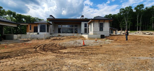 A wide exterior view of a newly constructed, low-profile contemporary home on a partially cleared and still-under-construction site. The house features pale masonry walls, dark metal hipped roof sections, extensive glazing, timber-clad elements, broad eaves, and a covered outdoor connection at the left. Bare earth, construction tracks, traffic cones, building materials, and a worker occupy the foreground and surroundings, with dense trees behind the property.